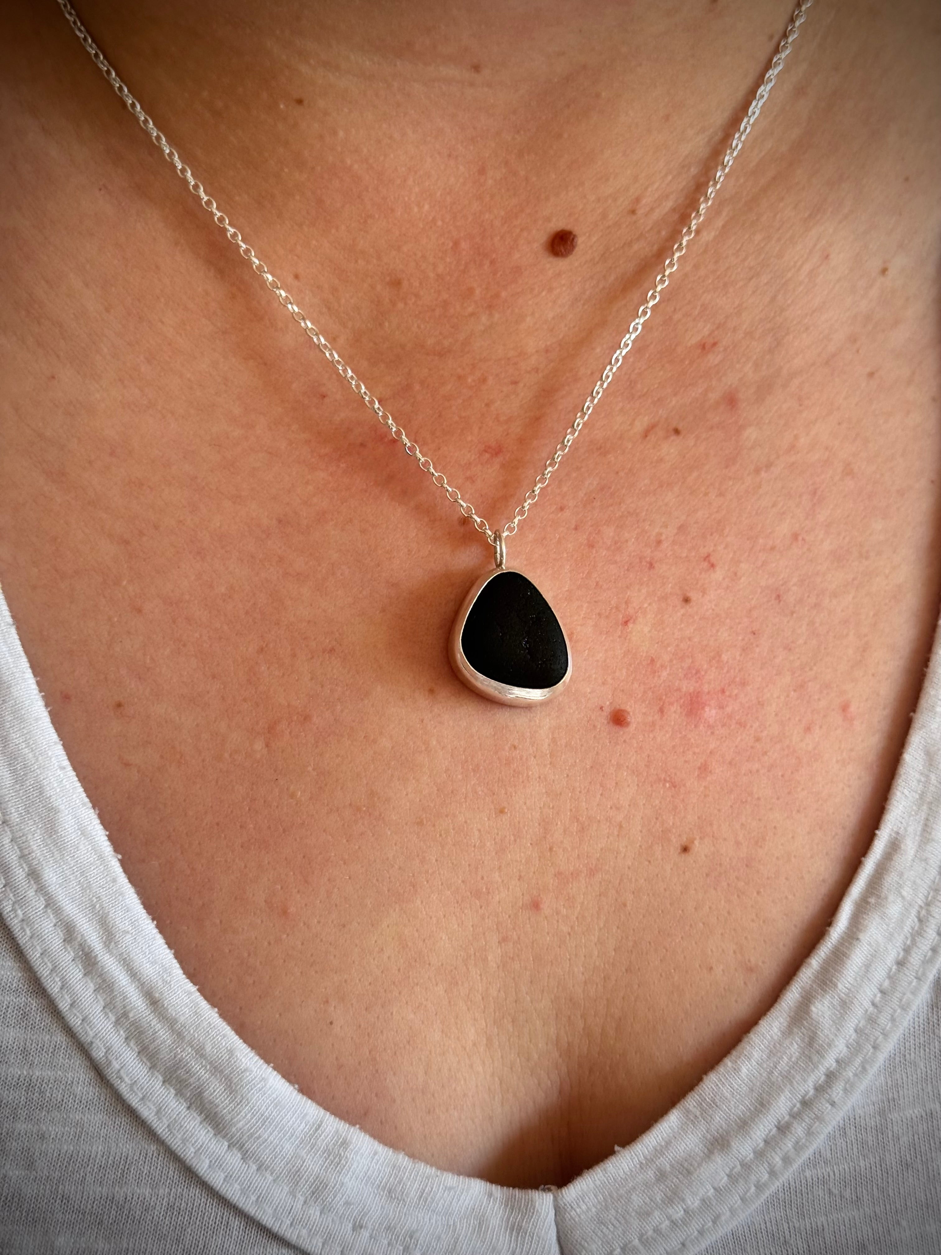 A close-up of a person's chest wearing a silver chain necklace with dark green sea glass pendant.
