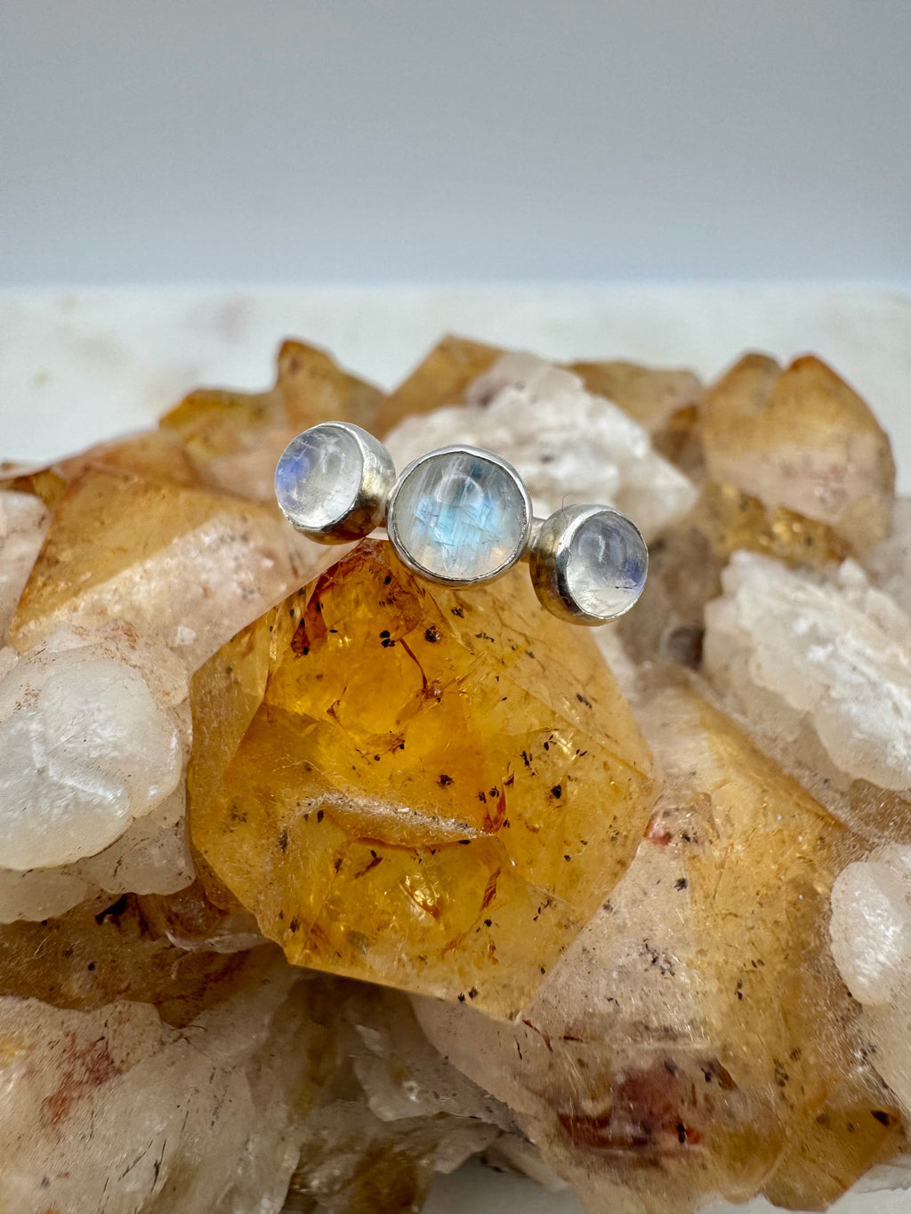 A sterling silver ring with three round-cut moonstone gemstones, displayed on top of a natural crystal cluster featuring a prominent golden citrine and white quartz.