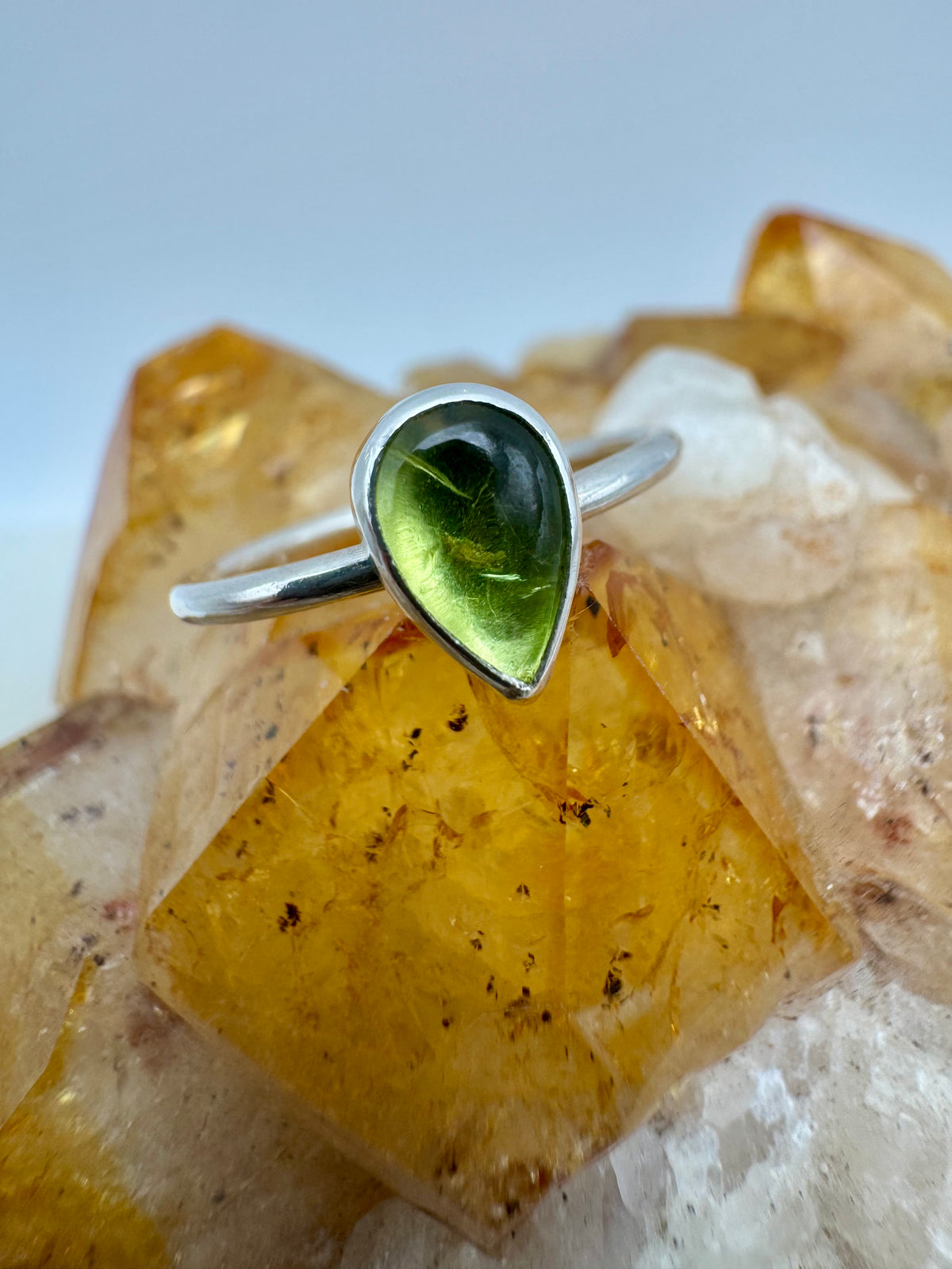 A sterling silver ring featuring a teardrop-shaped green peridot gemstone, displayed on a yellow crystal cluster against a light gray background.