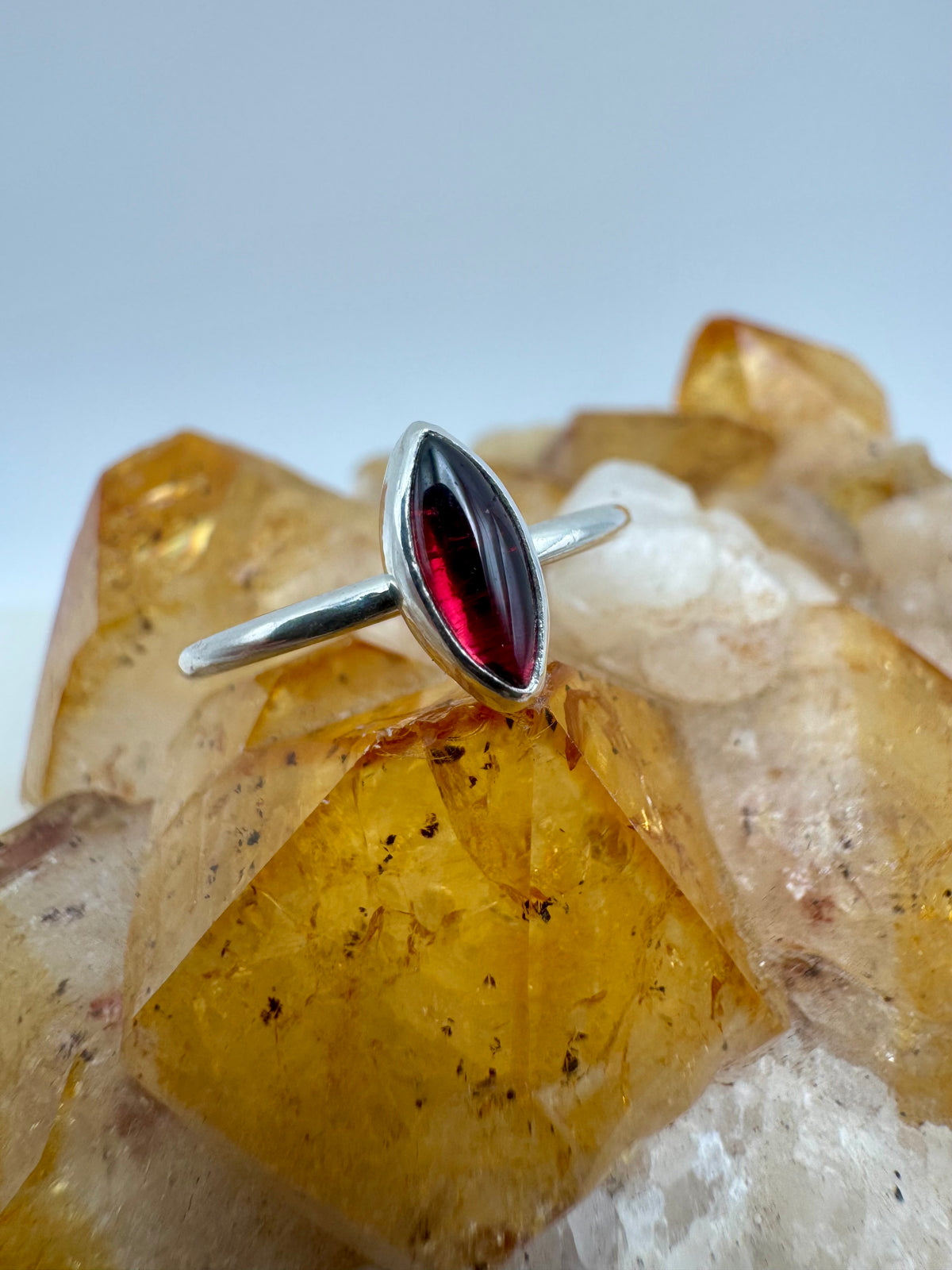 A silver ring with a marquise-cut dark red garnet gemstone, displayed on a cluster of yellow and white crystals against a light gray background.