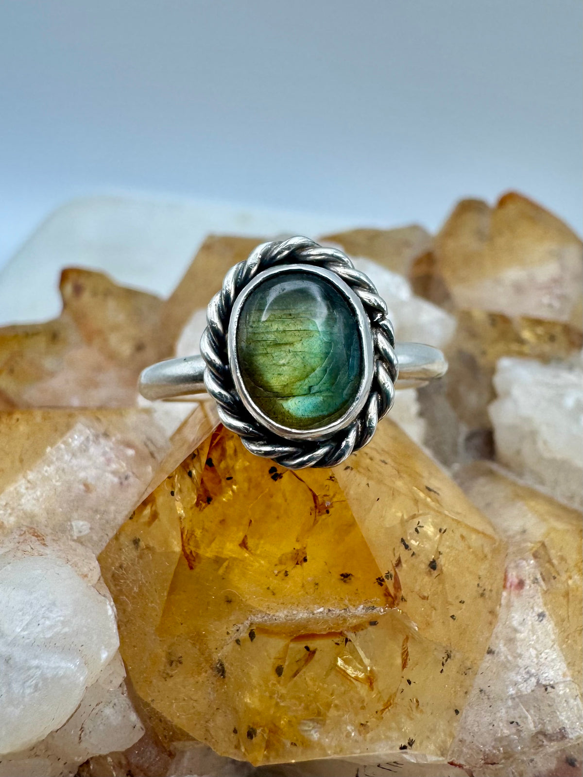 A silver ring with a twisted rope border and an oval green iridescent labradorite, displayed on a cluster of yellow and white crystals.