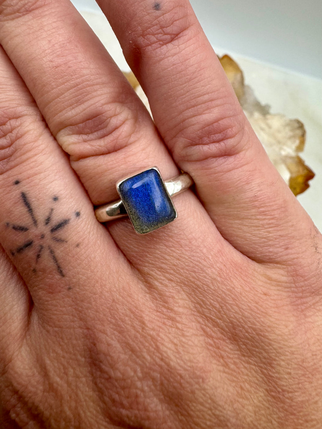 A close-up of a hand wearing a silver ring with a rectangular labradorite stone, set against a soft-focus background.