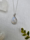 A silver teardrop pendant necklace with a moonstone, displayed on a textured white linen fabric with crystal and green leaf props.