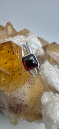 A silver split band ring with a square-cut dark red garnet gemstone, displayed on a crystal cluster with yellow and white stones against a light gray background.