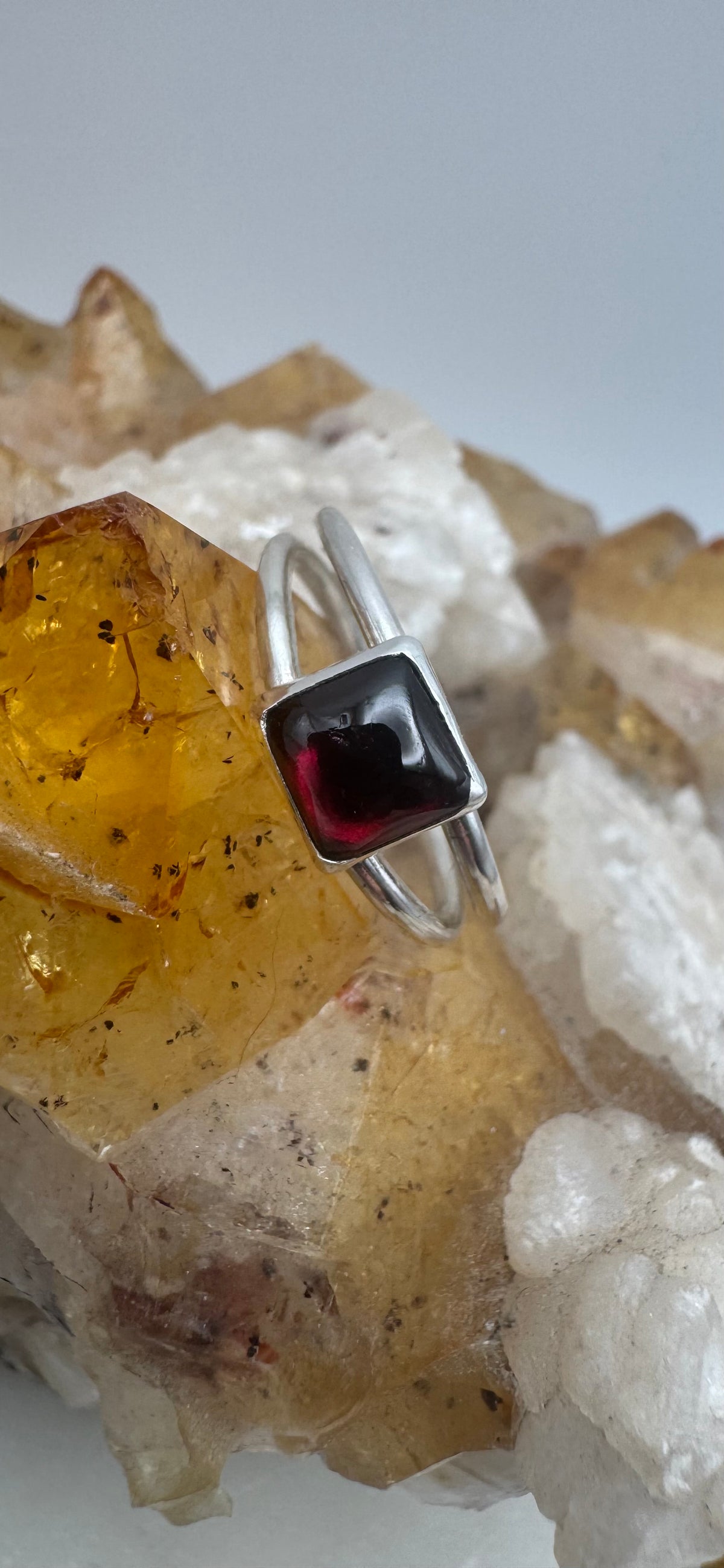 A silver split band ring with a square-cut dark red garnet gemstone, displayed on a crystal cluster with yellow and white stones against a light gray background.