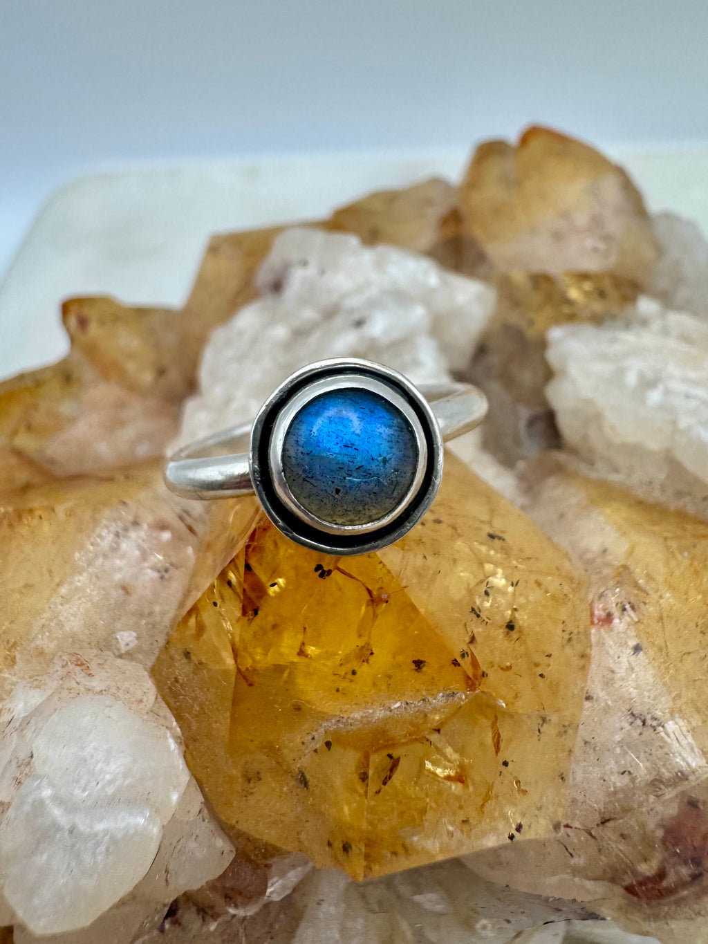 A sterling silver ring featuring a round bezel-set blue labradorite stone with a wire border,  on a cluster of natural quartz and citrine crystals.