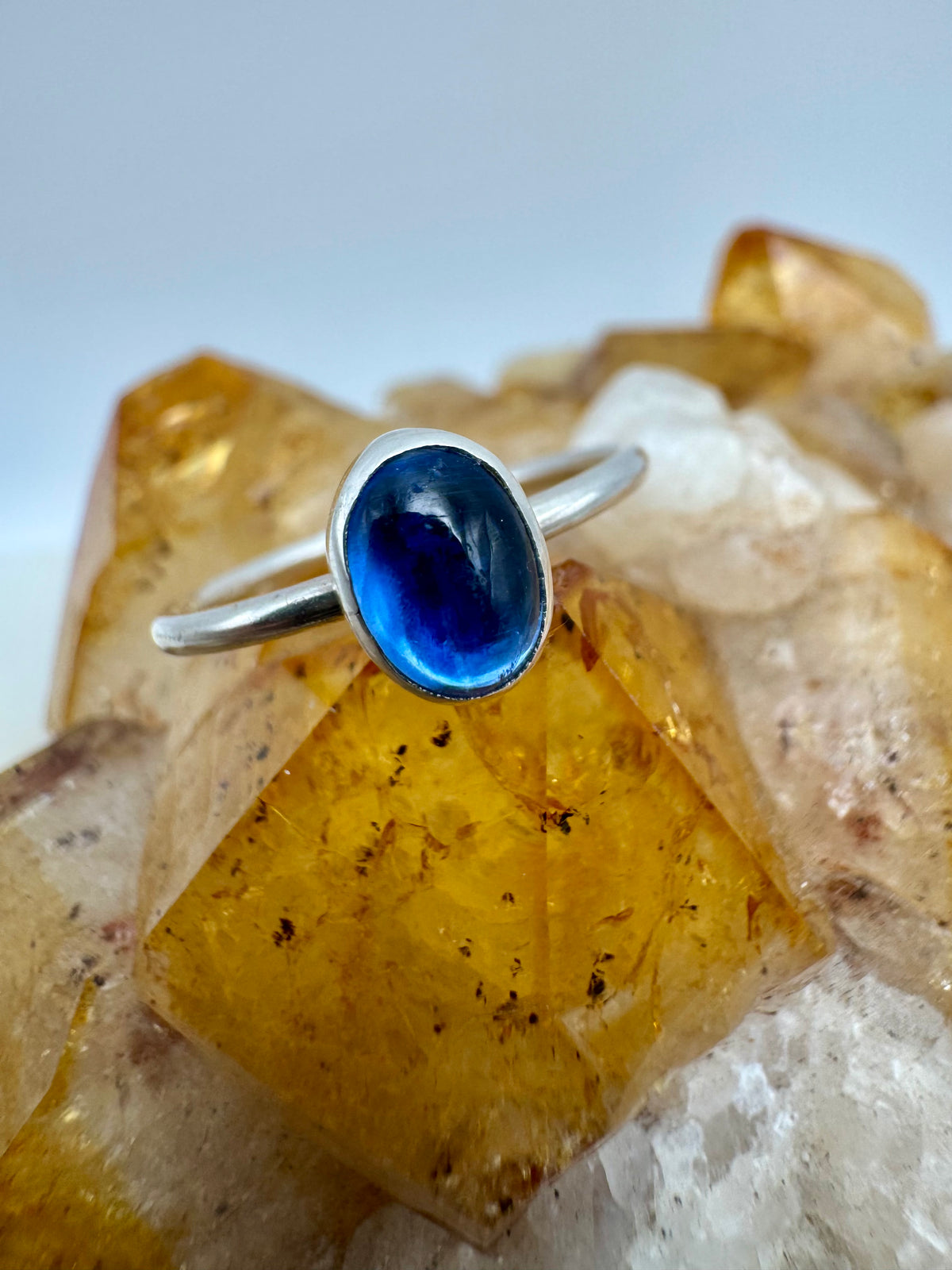A silver stacking ring with an oval blue iolite cabochon gemstone, displayed on a cluster of yellow and white crystals against a light blue-gray background.