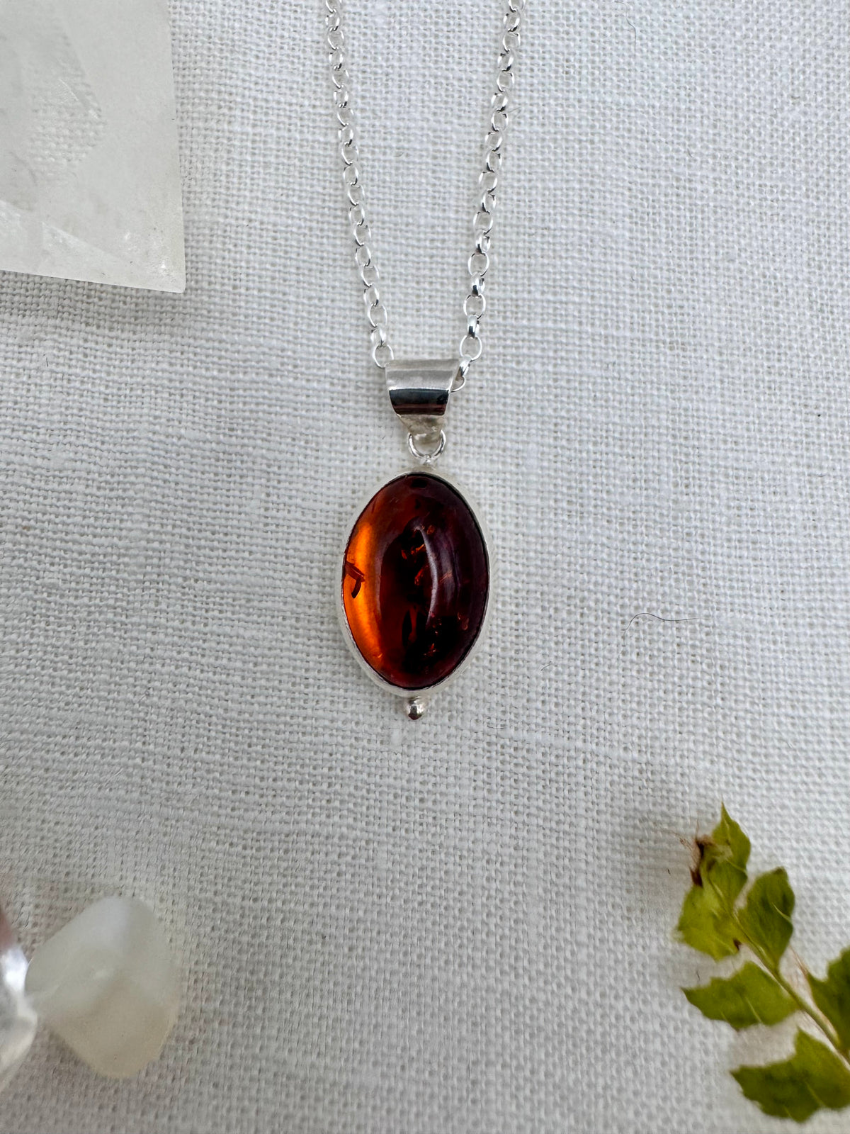 An oval amber pendant necklace with a small silver granule below, silver bezel and chain, displayed on a textured white fabric background with decorative crystals and a small green leaf.