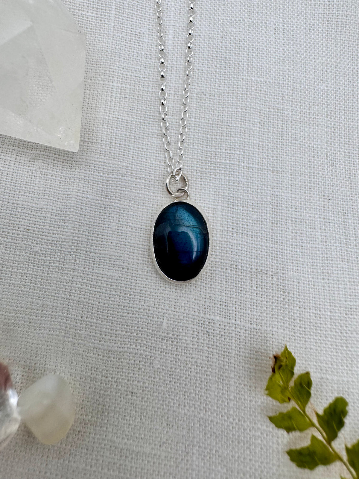 A silver chain necklace with an oval pendant featuring a deep blue labradorite, displayed on a textured white linen fabric with crystal and green leaf props.