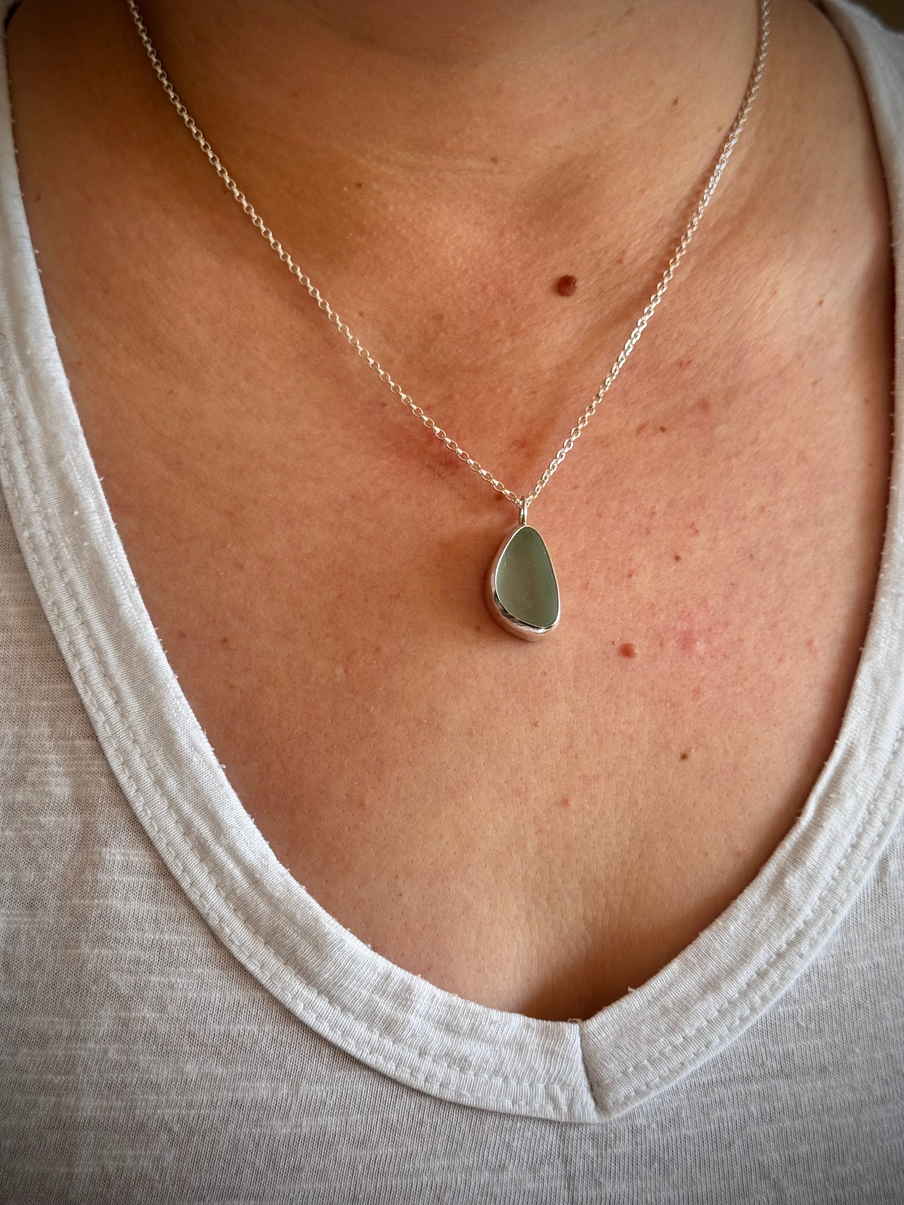 A close-up of a person wearing a delicate silver chain necklace with a teardrop-shaped sea glass pendant. The pendant is a muted sage green, set in a silver bezel, and is displayed against the wearer's chest.