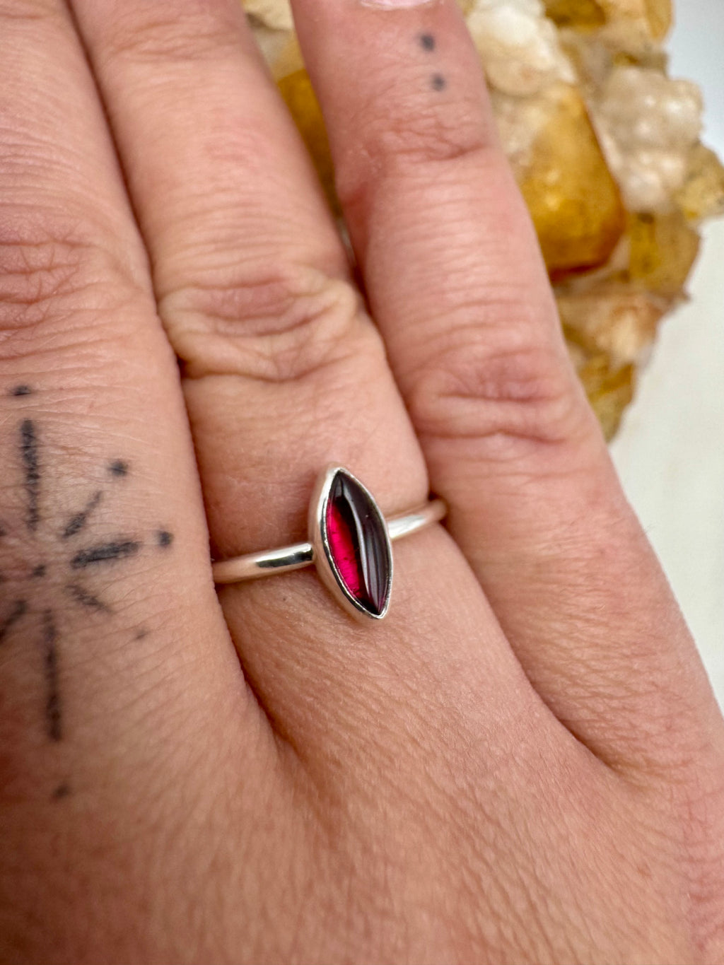A close-up of a hand wearing a silver stacking ring with a marquise-cut dark red garnet gemstone.