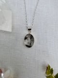 A silver chain necklace with an oval pendant featuring a black and white reclaimed quartz cabochon, displayed on a textured white fabric background with crystal props.