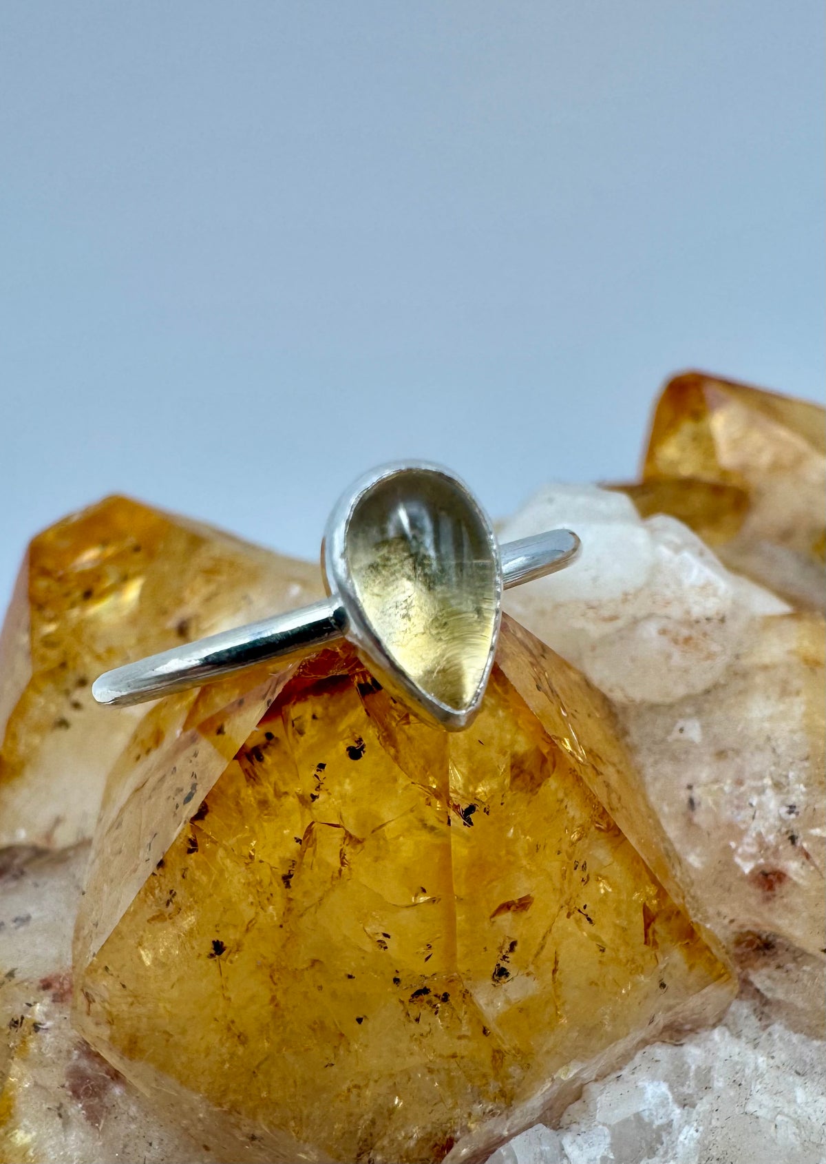 A silver ring with a teardrop-shaped yellow citrine gemstone, displayed on a cluster of yellow and white crystals against a light gray background.