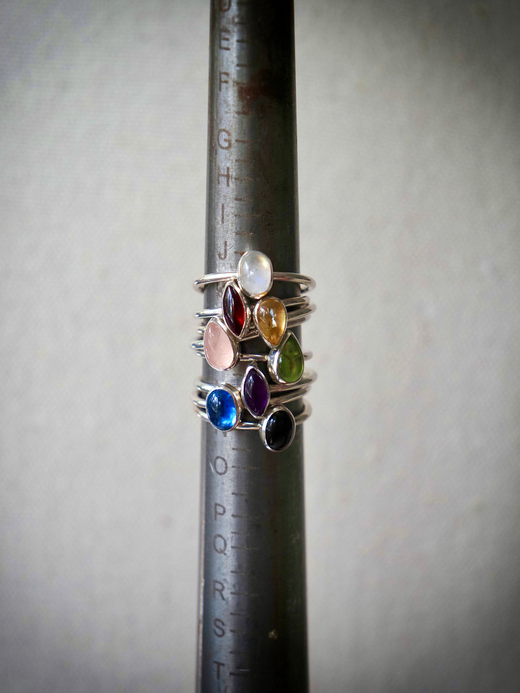 A stack of eight silver rings with colorful gemstones displayed on a dark metal ring sizer. The rings feature teardrop-shaped stones in white, red, pink, yellow, green, blue, and black.