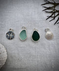 Four silver sea glass, seaweed and shell charms, displayed on a textured light gray fabric background with decorative seashells and dried seaweed.