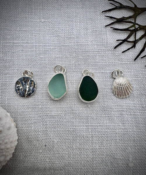 Four silver sea glass, seaweed and shell charms, displayed on a textured light gray fabric background with decorative seashells and dried seaweed.