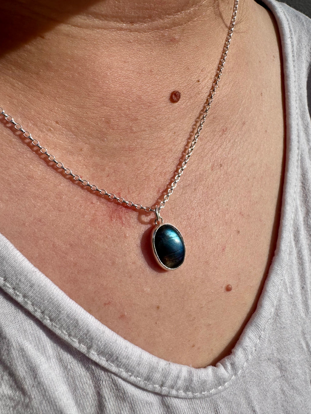A close-up of a person wearing a silver chain necklace with an oval dark blue iridescent labradorite pendant, styled with a light gray t-shirt.