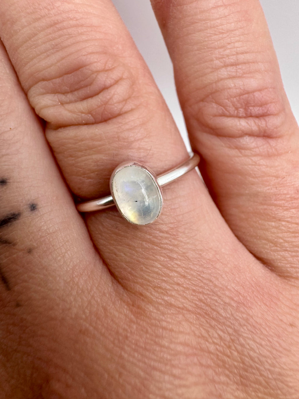 A silver ring with an oval, milky white cabochon gemstone featuring subtle iridescent rainbow flashes, shown worn on a person's finger.