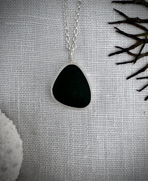 A silver pendant necklace featuring an irregular dark green sea glass piece, displayed on a textured white linen fabric with decorative dried seaweed and a white sea urchin.