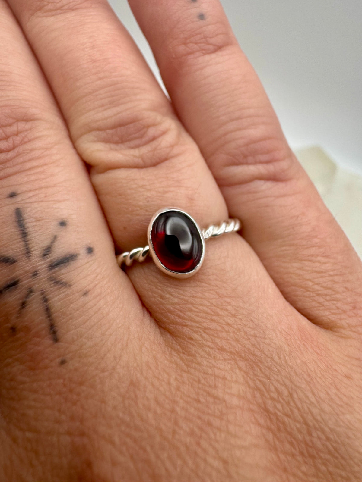 A silver ring with a twisted band and an oval garnet  cabochon gemstone worn on a person's finger, shown in a close-up shot.