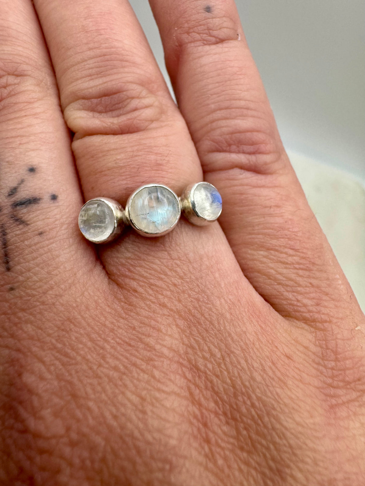 A close-up of a hand wearing a silver ring set with three round, iridescent moonstone cabochons.