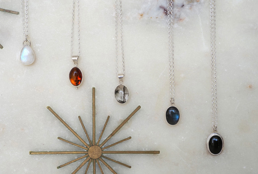 A set of five silver pendant necklaces with various gemstone drops, arranged on a light marble surface next to a gold sunburst star ornament. The necklaces feature moonstone, amber, quartz, labradorite, and onyx stones.