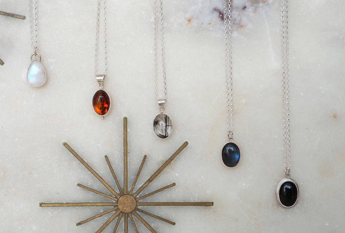 A set of five silver pendant necklaces with various gemstone drops, arranged on a light marble surface next to a gold sunburst star ornament. The necklaces feature moonstone, amber, quartz, labradorite, and onyx stones.