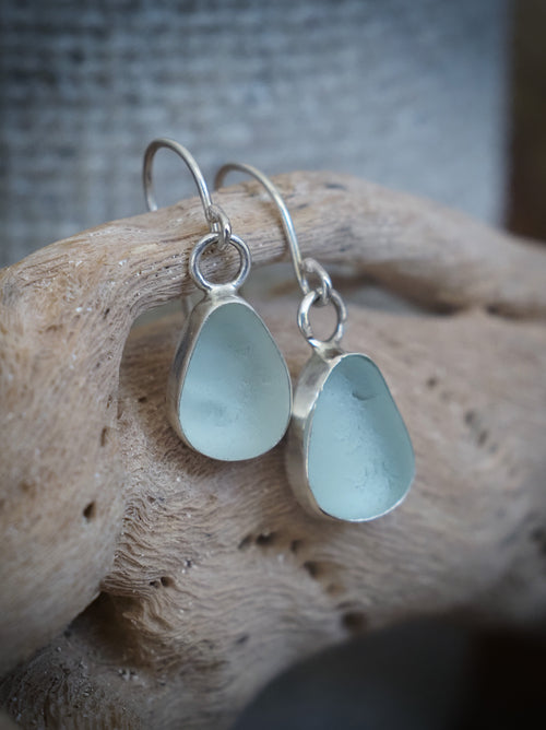 A pair of silver drop earrings with teardrop-shaped sea glass pendants in a seafoam aqua hue, displayed on a piece of driftwood.