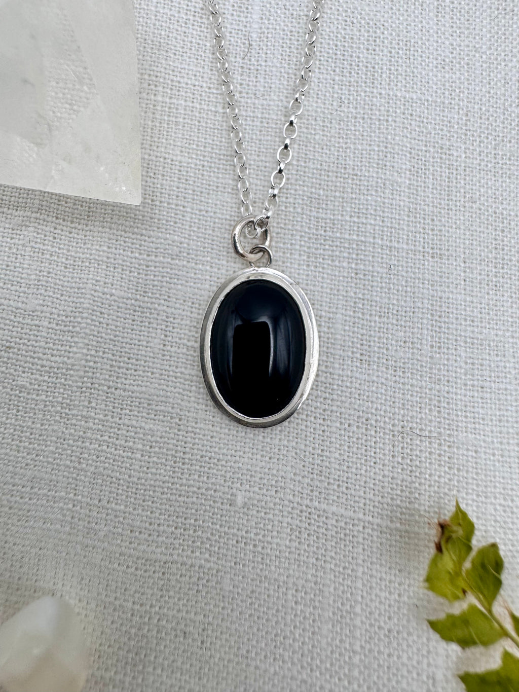 A silver oval pendant necklace with an onyx stone and silver border, displayed on a textured white fabric background with a crystal and green leaves as props.