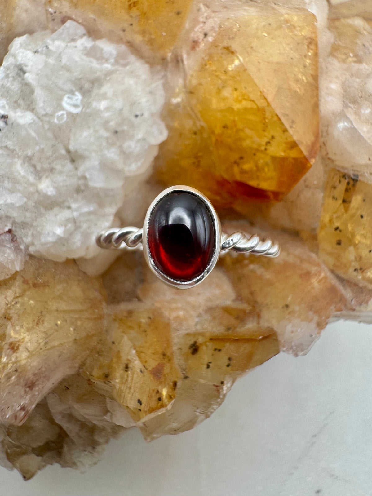 A silver ring with a twisted band and garnet oval gemstone, displayed on a natural crystal cluster.