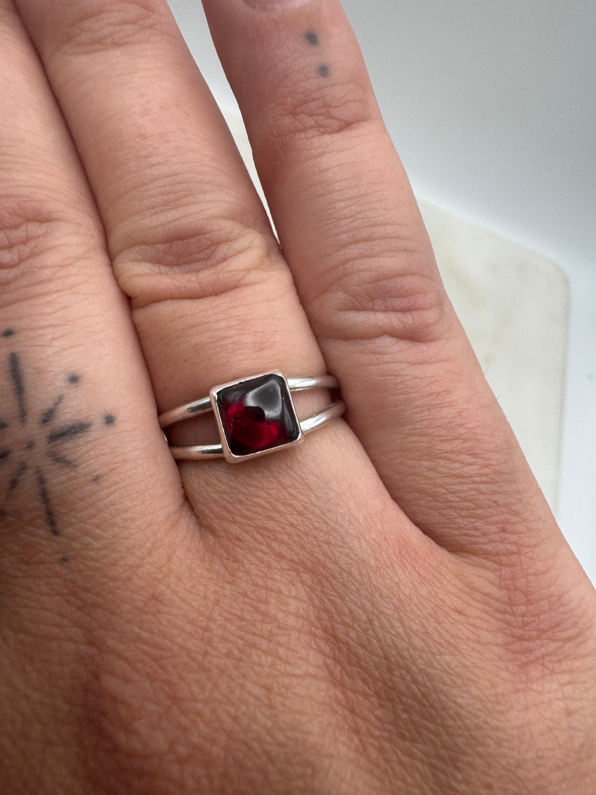 A close-up of a hand wearing a silver double-band ring with a square-cut dark red garnet gemstone.
