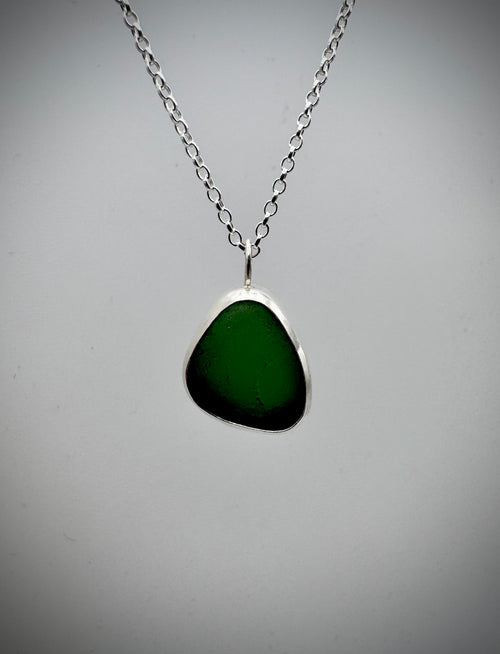 A silver chain necklace with an irregular green sea glass pendant set in a silver bezel, open backed with the light shining through, shown against a neutral gray background.