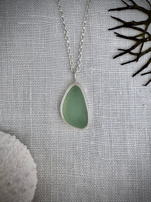 A silver necklace with an irregular teardrop-shaped sea glass pendant in a light green shade, displayed on a textured white linen background with decorative seashells and dark coral.
