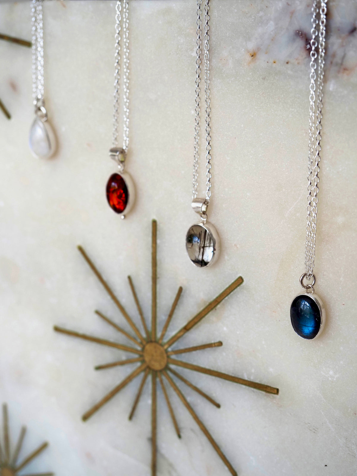 Four silver chain necklaces with oval and teardrop pendants displayed against a light marble-like surface with a gold starburst decoration. The pendants include moonstone, quartz and labradorite stones.