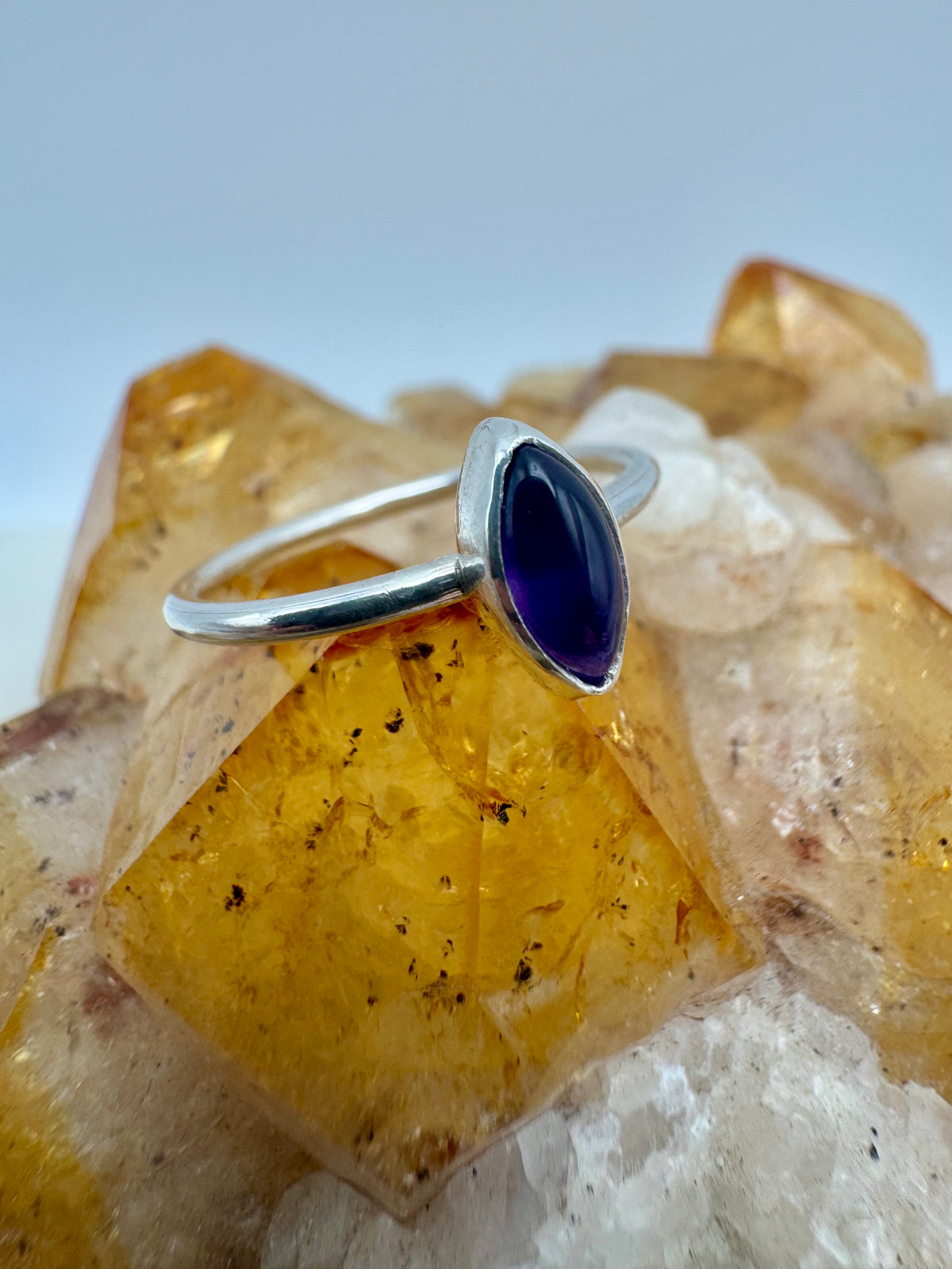 A silver stacking ring with a marquise-cut dark purple amethyst gemstone, displayed on a yellow and white crystal cluster.