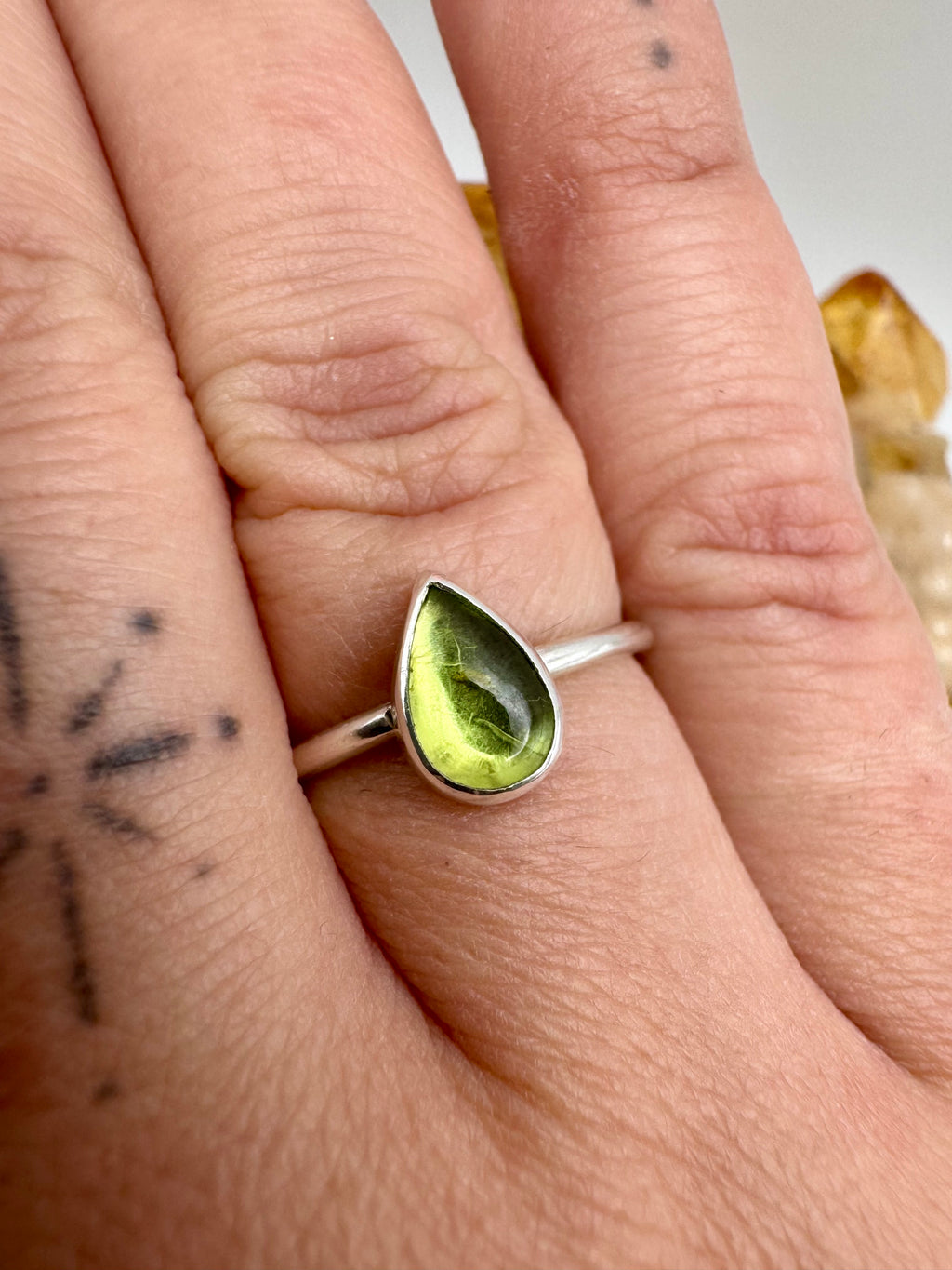A silver ring with a teardrop-shaped green peridot gemstone worn on a person's finger, shown in a close-up shot.