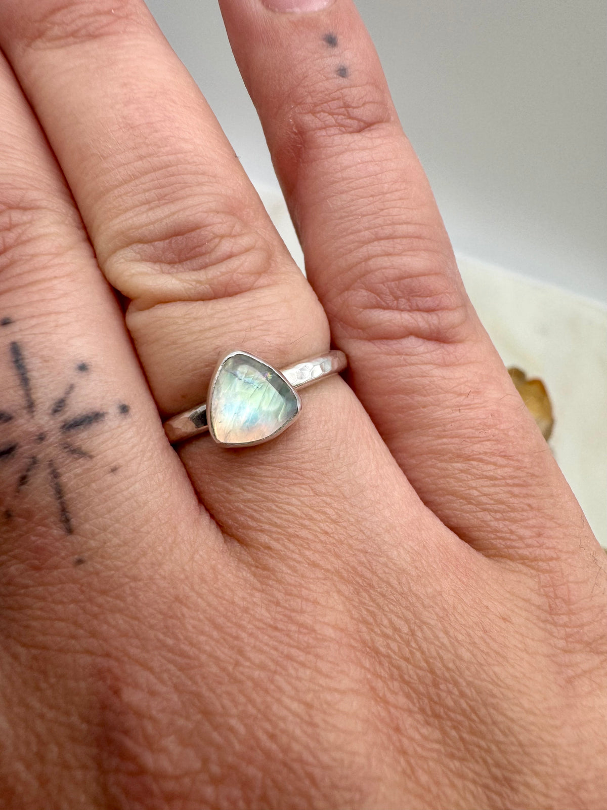 A silver ring with a triangular moonstone worn on a person's finger, shown in a close-up shot of the hand.