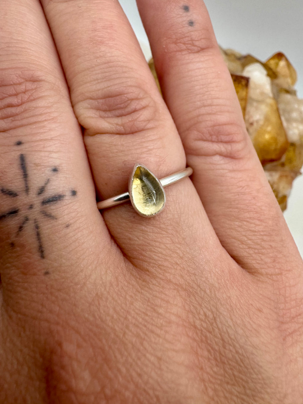 A close-up of a hand wearing a silver ring with a teardrop-shaped yellow citrine gemstone, with a crystal cluster visible in the background.