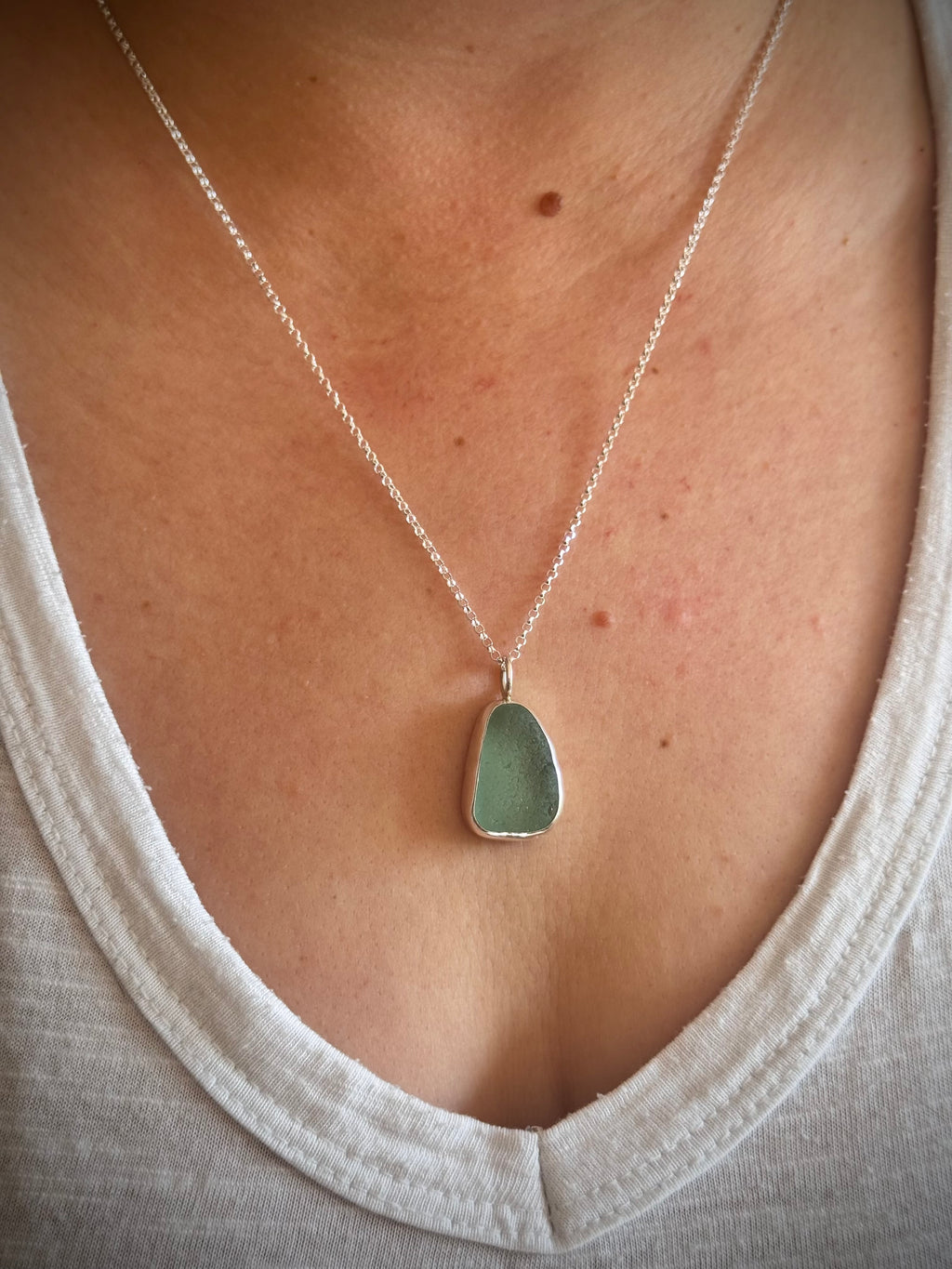 IA close-up of a person wearing a silver chain necklace with a smooth, irregular light green aqua pendant, styled with a white top.