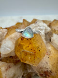 A silver ring with a triangular moonstone resting on a cluster of crystals, including a prominent yellow-orange quartz crystal.
