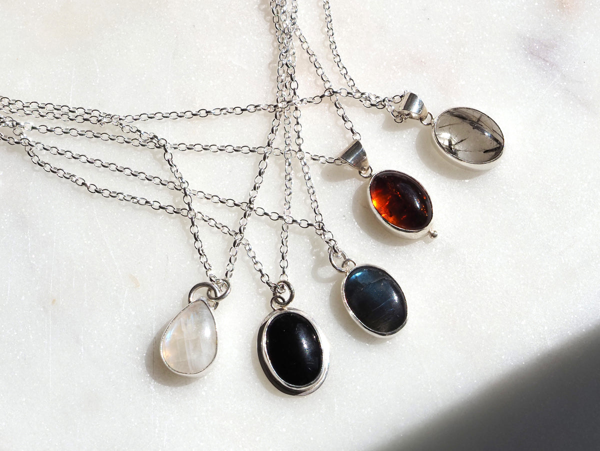 Five silver chain necklaces with teardrop and oval pendants in different coloured stones, arranged on a light marble surface.
