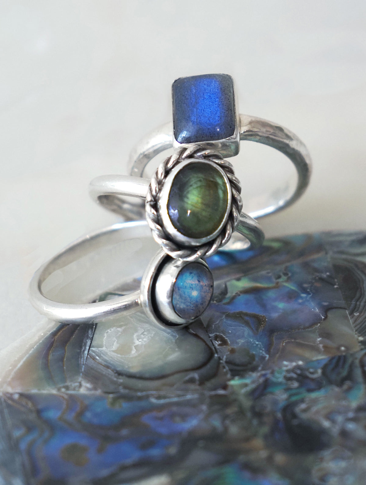 A silver ring set featuring a square-cut blue stone, an oval green labradorite stone, and a smaller round blue stone, displayed on a piece of abalone shell.