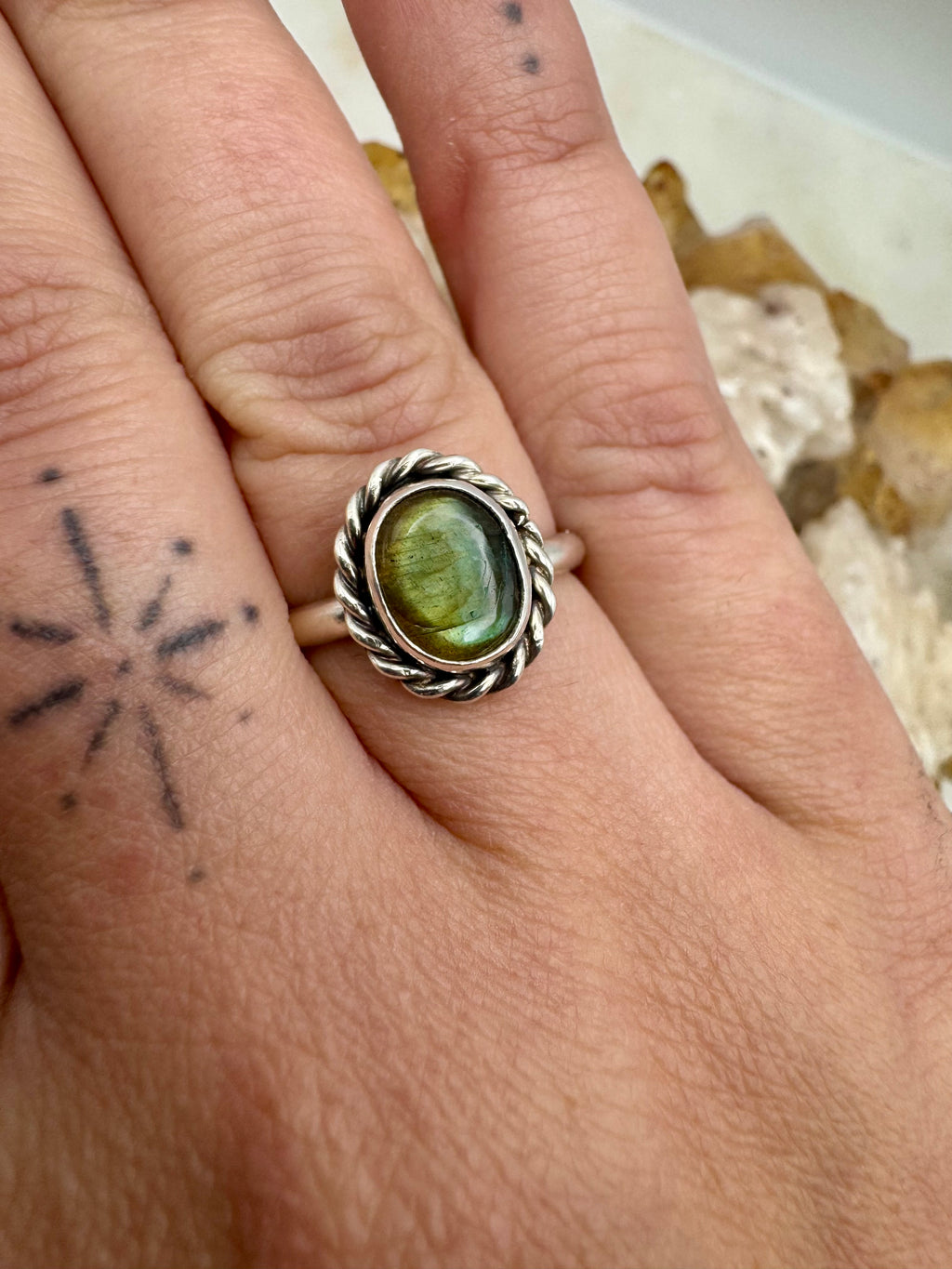 A close-up of a hand wearing a silver ring with an oval labradorite cabochon stone. The ring features a twisted rope border and is shown on the middle finger against a neutral, slightly blurred background.