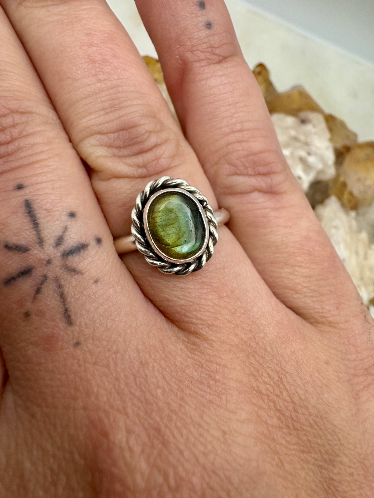 A close-up of a hand wearing a silver ring with an oval labradorite cabochon stone. The ring features a twisted rope border and is shown on the middle finger against a neutral, slightly blurred background.