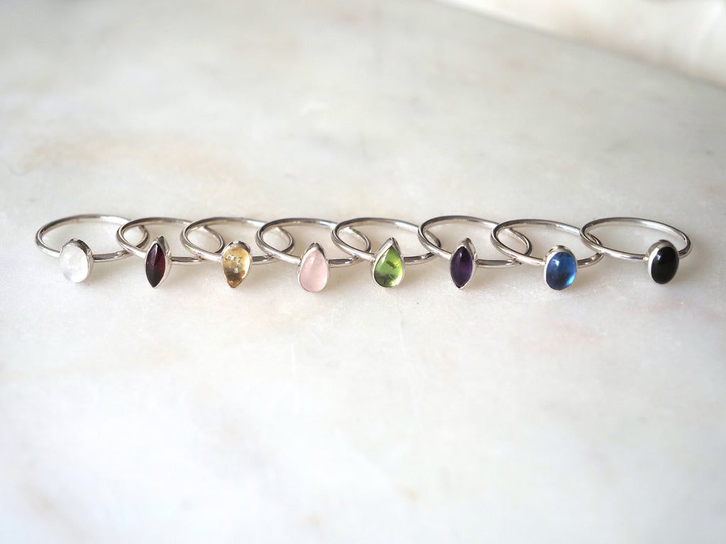 Eight silver stackingrings with teardrop and oval shaped gemstones in various colours, arranged in a row on a light marble surface.