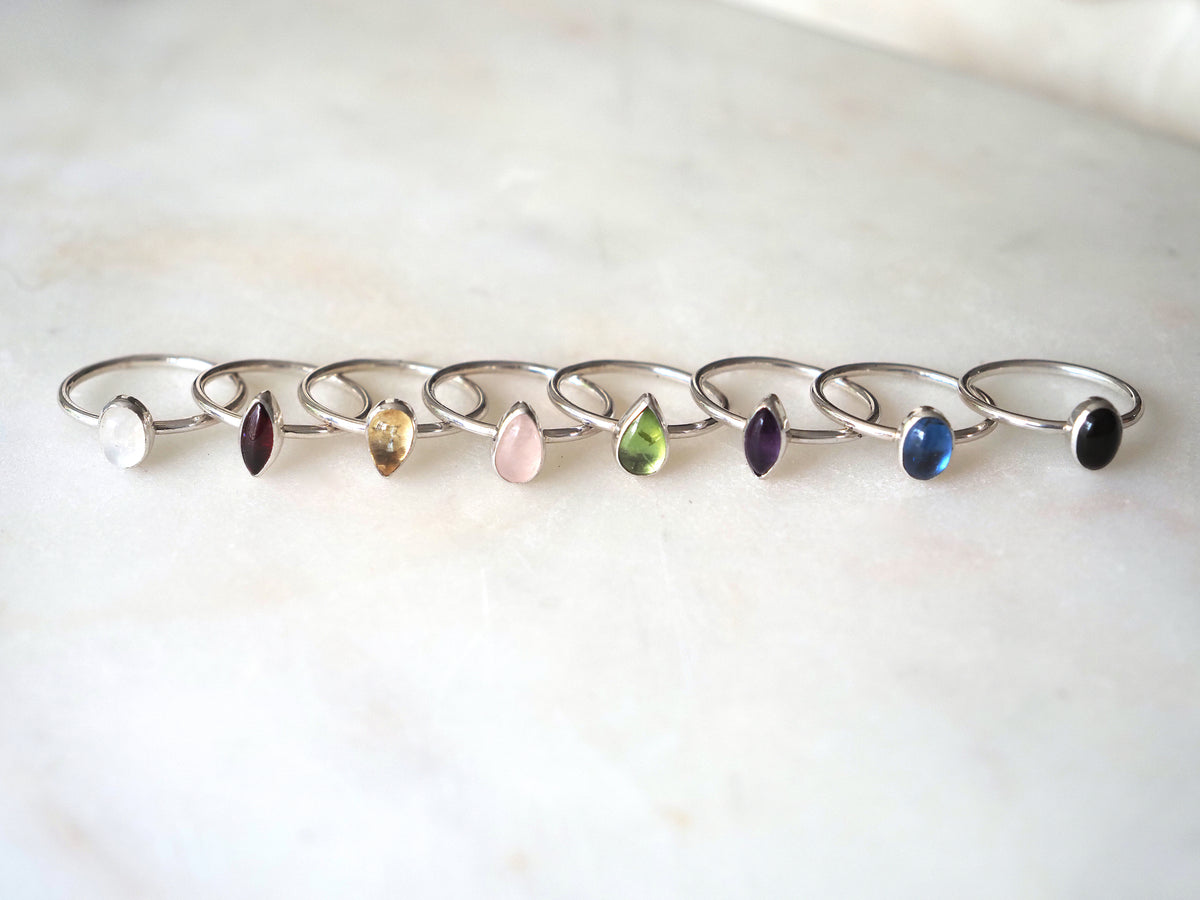 Eight silver stackingrings with teardrop and oval shaped gemstones in various colours, arranged in a row on a light marble surface.
