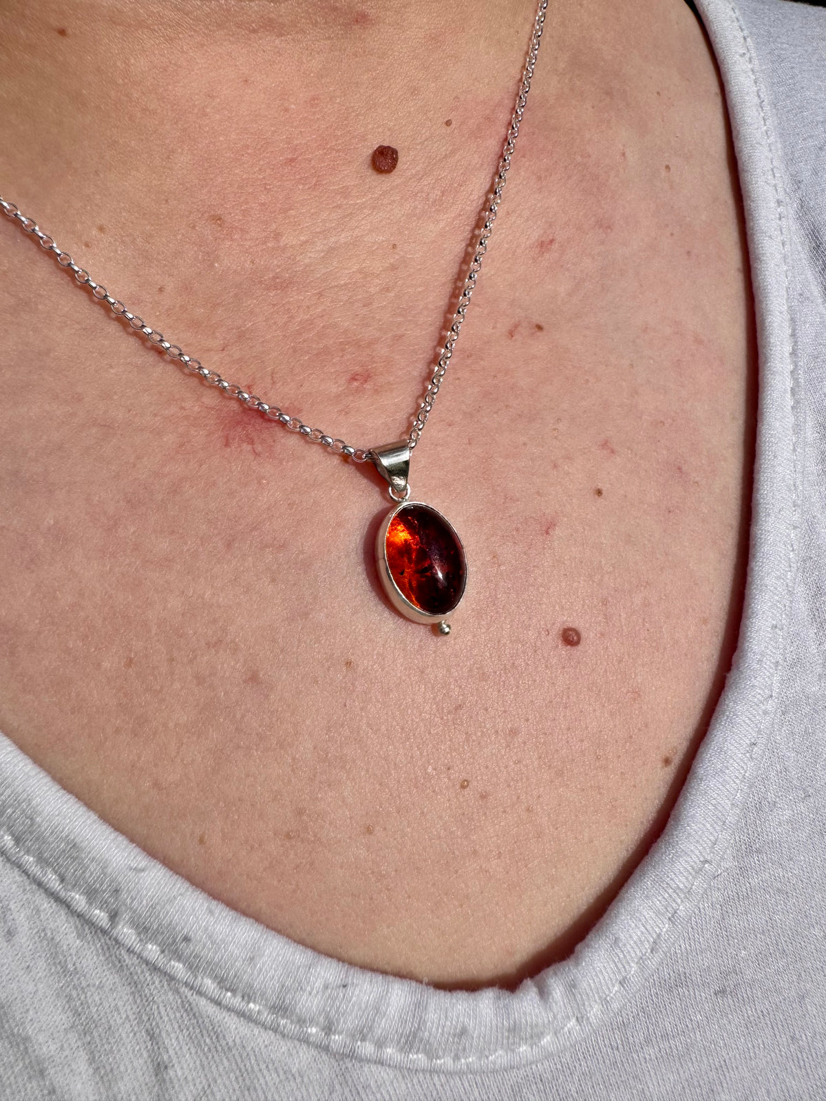 A close-up of a person wearing a silver chain necklace with an oval amber gemstone pendant, shown against the chest and shoulders.