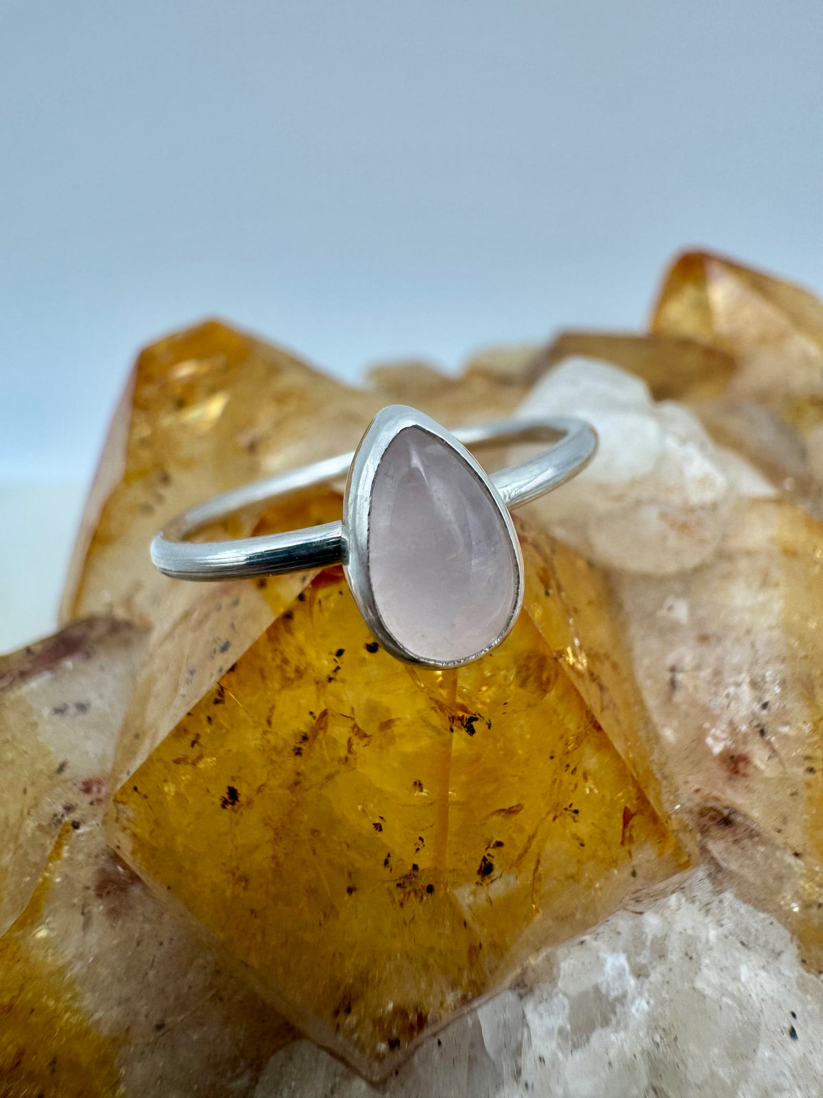 A silver ring with a teardrop-shaped pale pink rose quartz stone, displayed on a cluster of yellow and white crystals against a light gray background.