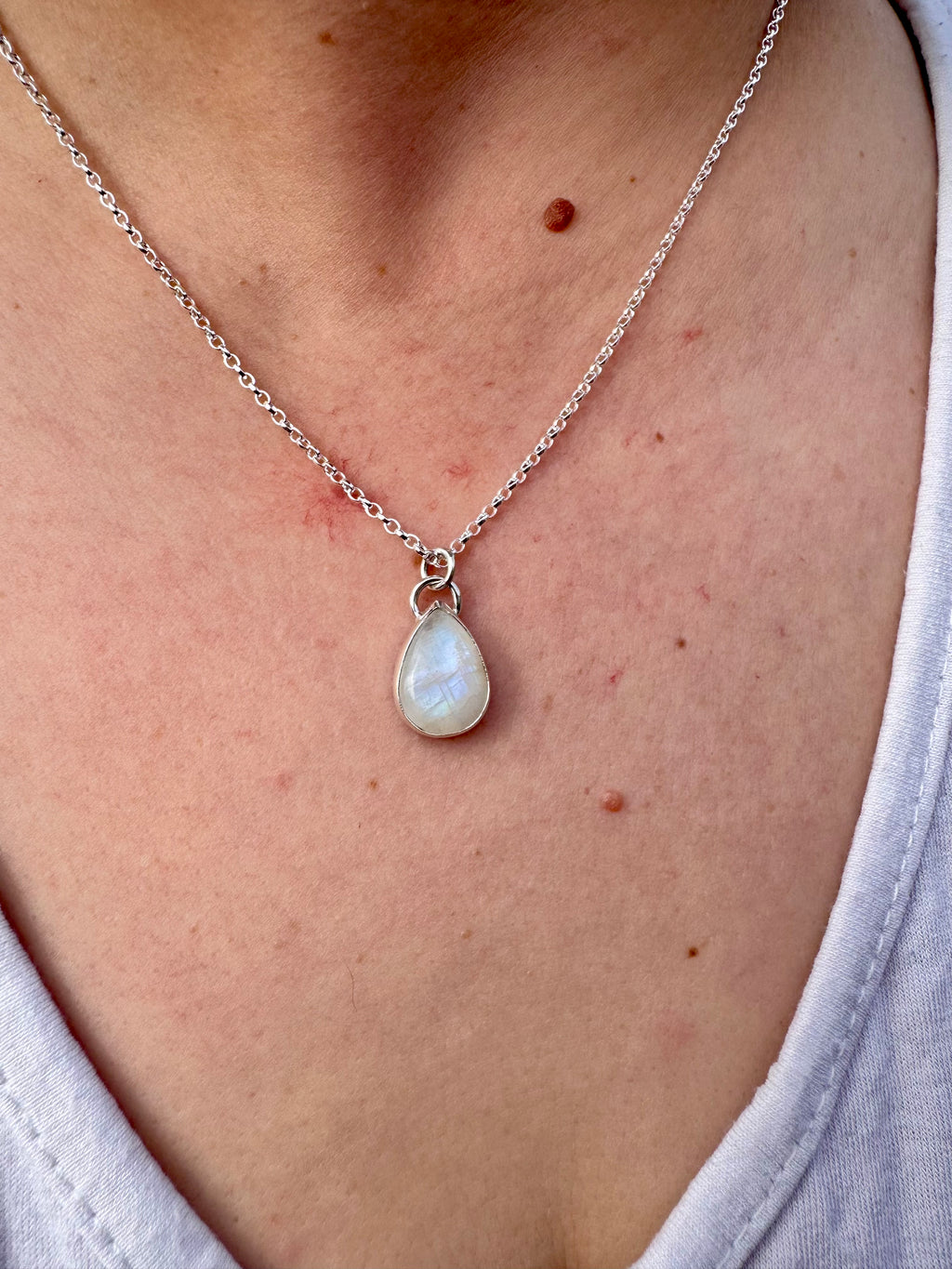 A close-up of a person's neck and chest wearing a delicate silver chain necklace with a teardrop shaped moonstone pendant.