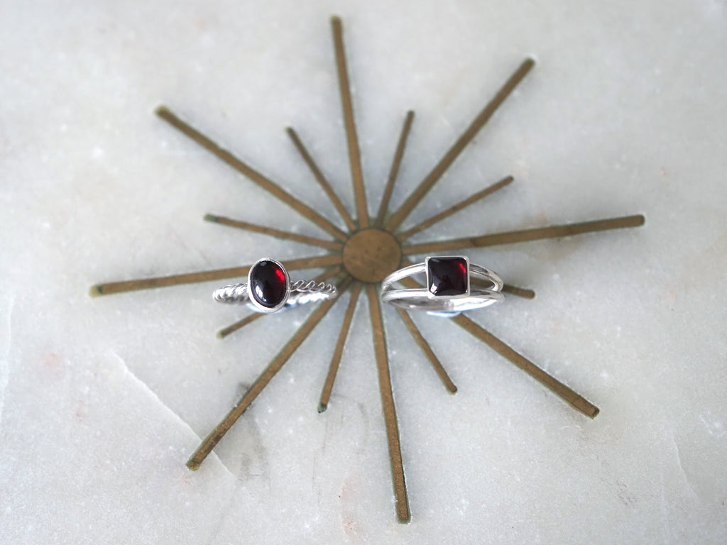 Two silver rings with garnet gemstones displayed on a brown starburst stand against a light marble background.